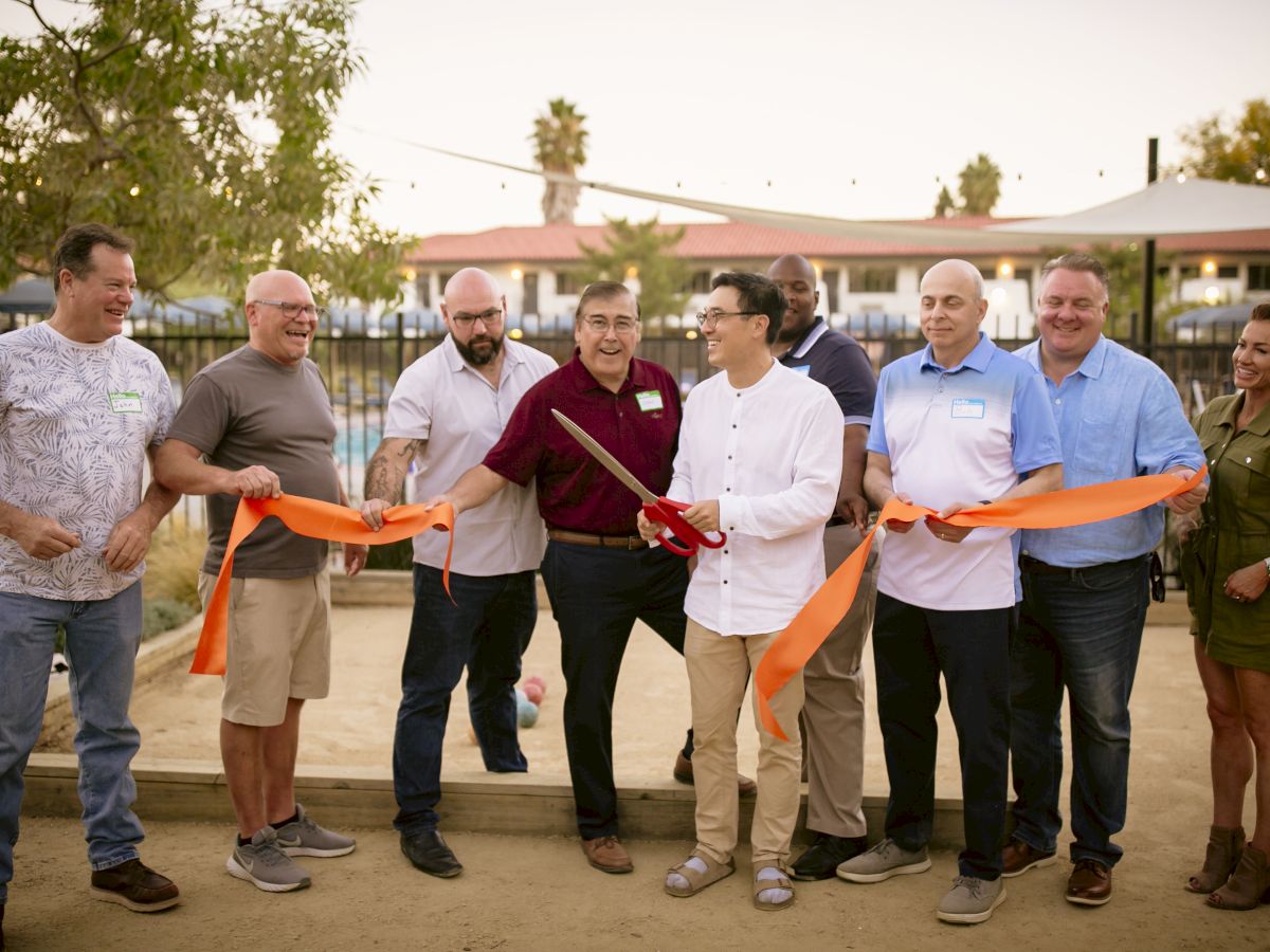 A group of people stands together outdoors, holding an orange ribbon and a pair of large scissors, presumably for a ribbon-cutting ceremony, ending the sentence.