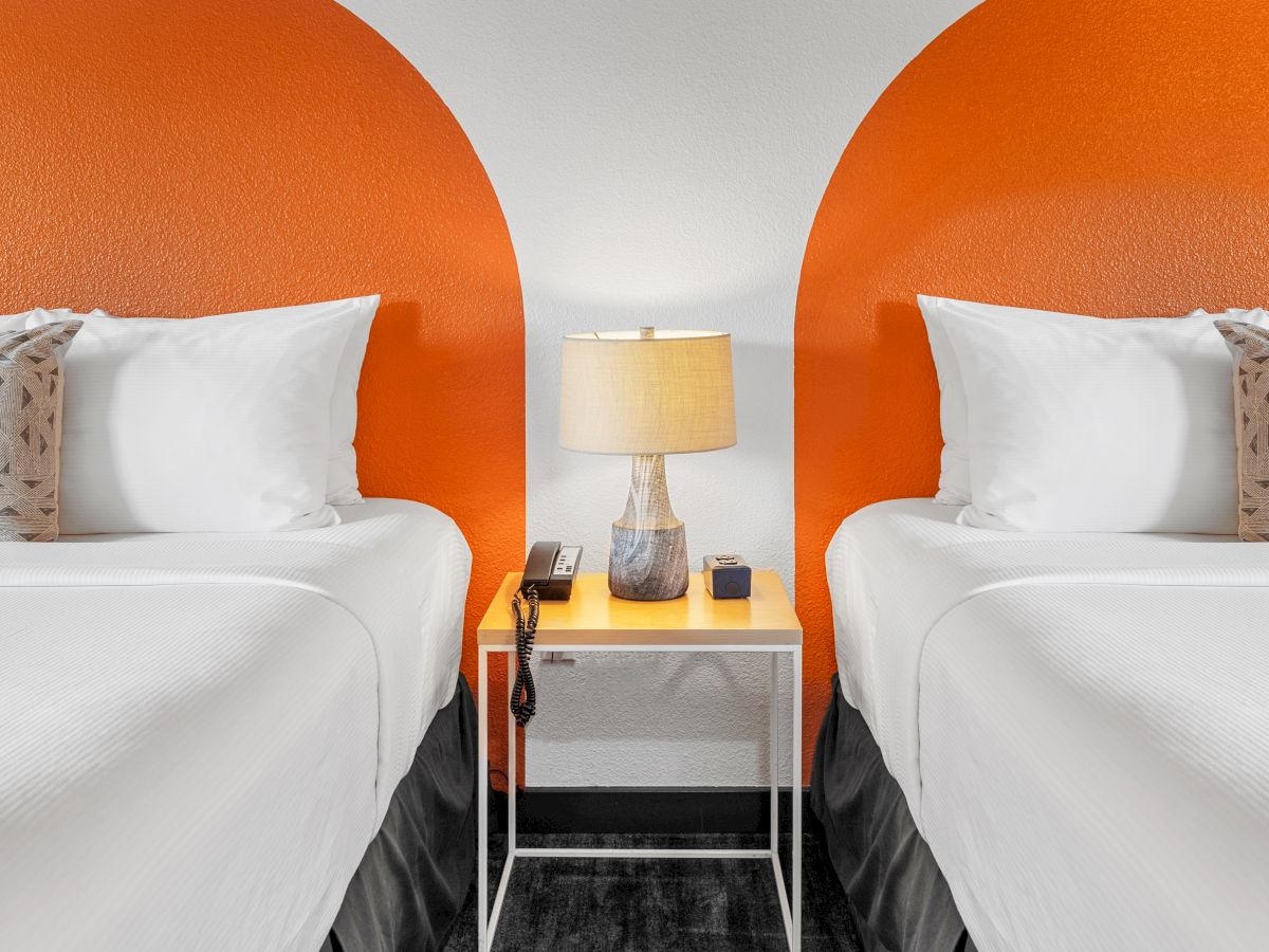 The image shows two white beds with pillows, separated by a small nightstand with a lamp, phone, and decor. Orange semicircles are painted on the wall.